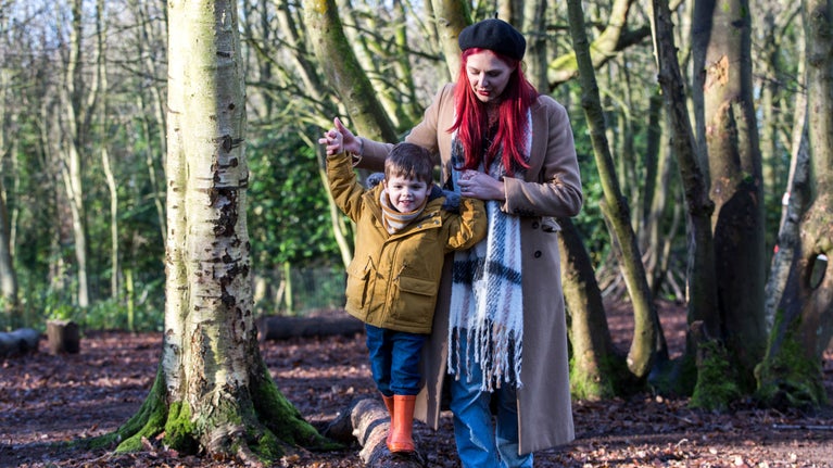 A mother holds her sons hands above his head, as he balances on a log beam in a wintry woodland